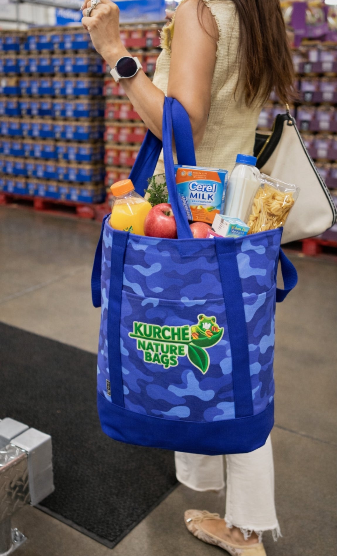 Woman carrying a Nautical Blue Camo Kürché Nature Bags reusable canvas tote bag filled with groceries in a supermarket aisle.