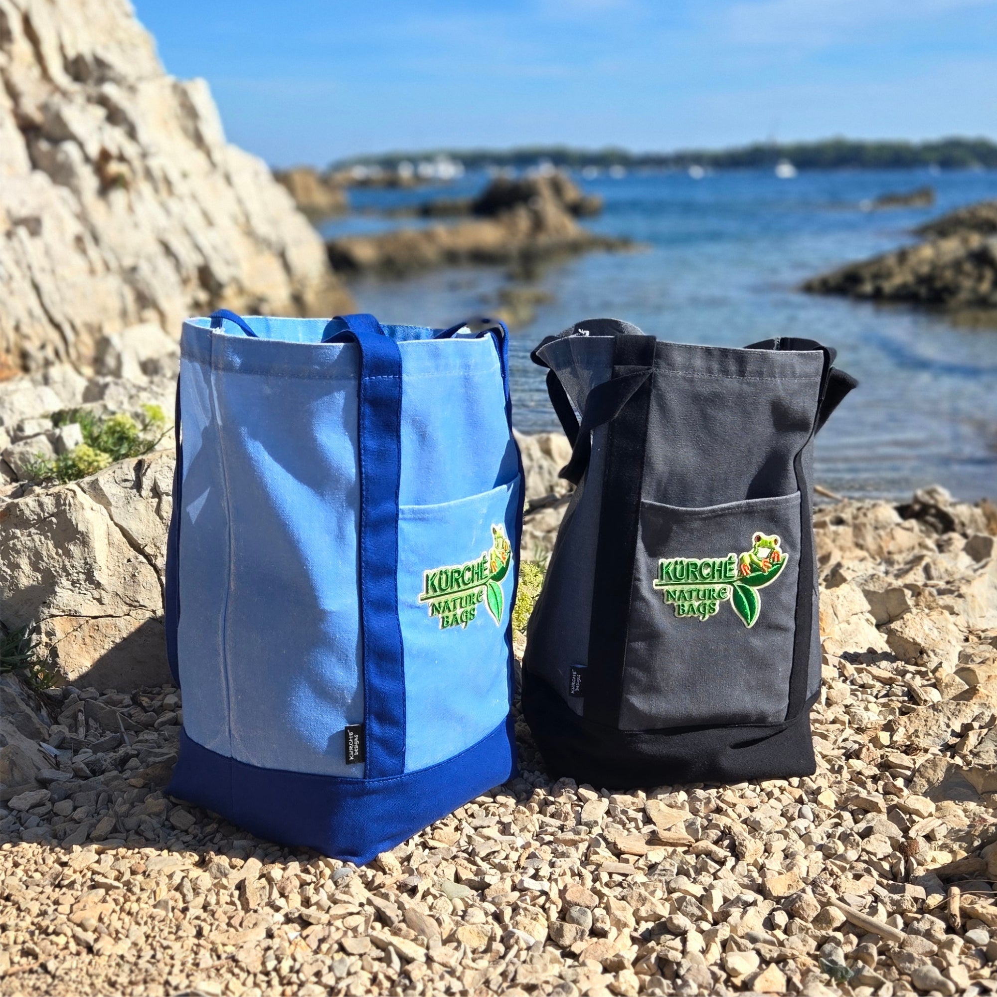 Blue and black Kürché Nature Bags reusable canvas tote bags displayed on a rocky beach by the water