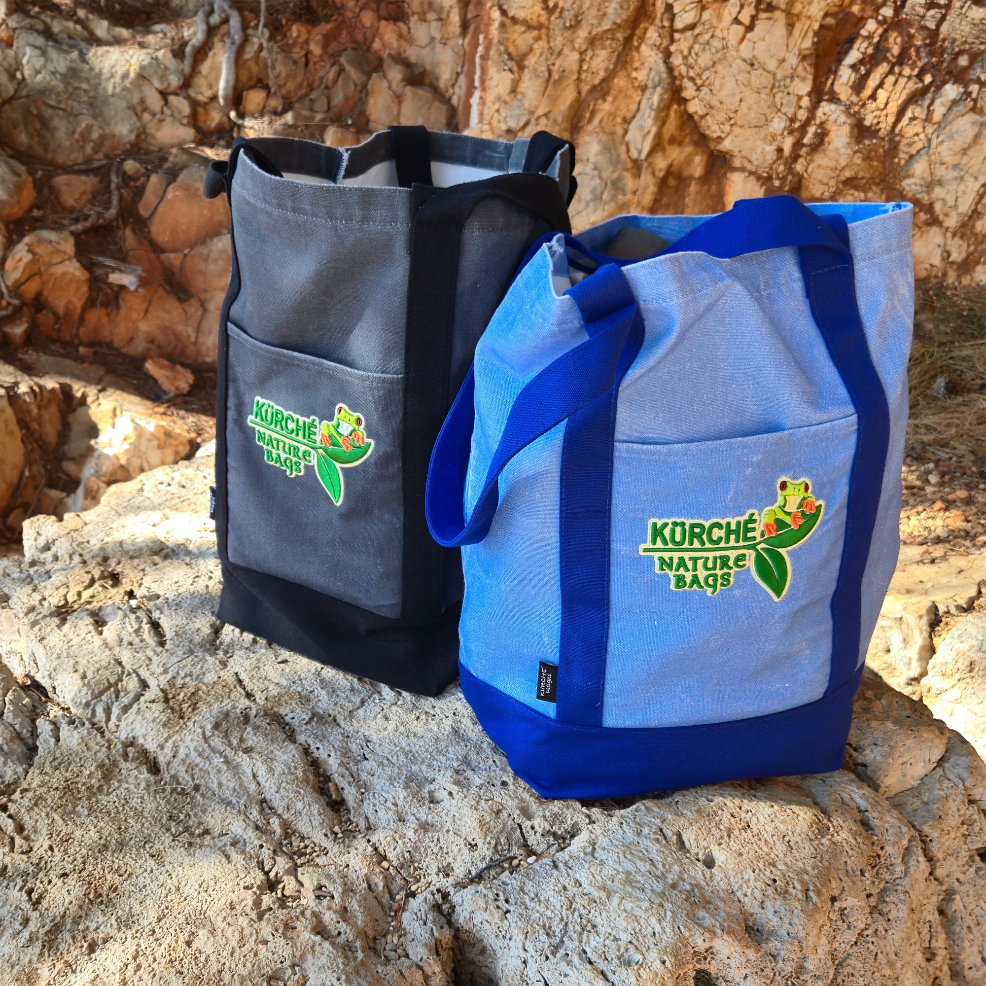 Blue and midnight black reusable canvas tote bags with embroidered KÜRCHE Nature Bags logo displayed outdoors on rocks