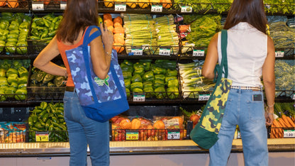 Two women examining fresh vegetables in a store, each carrying a camo Kurche Nature Bag, highlighting versatility for shared shopping trips.